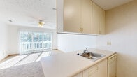 This single-floor unit presents a practical and well-organized layout with a neutral color palette throughout. The kitchen is efficiently designed, featuring light beige cabinetry extending to the ceiling for generous storage, a stainless steel sink set into a continuous light-toned countertop, a dishwasher, and a range. Tile flooring grounds the kitchen space, which opens toward the living area. The living area is carpeted and benefits from abundant natural light streaming through large sliding glass doors that lead to a balcony, offering pleasant outdoor views. A ceiling fan overhead adds both functionality and a sense of comfort to the space. The bedroom areas are also carpeted and feature textured ceilings, with one room equipped with track lighting and a window fitted with vertical blinds that allow natural light to filter through. Closet space is accessible via light wood-toned bifold doors, with additional storage visible nearby. The bathroom is accessed through a short hallway and includes a vanity with white cabinetry, a mirror, a combination tub and shower, and tile flooring. Light wood trim and door hardware in warm tones carry consistently through the unit, creating a cohesive and understated interior throughout.
