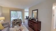 This single-floor residence offers a thoughtfully designed layout that balances comfort and functionality. The living area serves as the heart of the home, bathed in natural light streaming through large vertical-blind windows that frame lush outdoor greenery, creating a warm and welcoming atmosphere. Hardwood-style flooring runs throughout, adding warmth and a cohesive aesthetic to the space.

The living room is generously proportioned, providing ample room for relaxed seating arrangements, and flows seamlessly into an adjacent dining area, also brightened by its own set of windows. This open connection between spaces enhances the overall sense of openness and ease of movement throughout the home.

Neutral wall tones provide a versatile backdrop, complementing both the rich, dark wood tones of the furnishings and the light, airy feel of the interiors. A textured ceiling adds subtle architectural character. The space is styled with tasteful décor, including a bold-framed mirror, decorative greenery, and curated accents that highlight the home's inviting character. Overall, this residence offers a bright, comfortable, and well-connected living environment suitable for a variety of lifestyles.