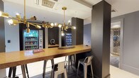 This single-floor space presents a polished, contemporary aesthetic designed for casual comfort and convenience. The interior is anchored by a warm-toned granite or stone bar counter with industrial-style metal bar stools, creating an inviting spot for guests to gather. A striking brass multi-arm pendant chandelier serves as the visual centerpiece, casting a warm ambient glow that contrasts elegantly with the space's dark charcoal accent walls and white ceiling. Recessed ceiling-mounted HVAC units and a flush white ceiling contribute to a clean, streamlined overhead finish. Smart home or building controls are integrated directly into the walls, reflecting a modern, tech-forward design approach. Along the perimeter, a well-stocked beverage refrigerator and a snack vending machine offer convenient self-service options, suggesting this space functions as a hotel lobby lounge, co-working amenity area, or hospitality suite. The dark slate-toned flooring ties the design together, reinforcing the sophisticated, urban character of the space. Overall, the layout is compact yet thoughtfully appointed, balancing style with practical functionality for everyday use.