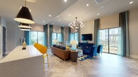 Located at 240 Park Place Blvd in Waxahachie, TX, this single-floor community clubhouse is designed with a bold, contemporary aesthetic that blends comfort and style. The main lounge area is the centerpiece of the space, boasting soaring ceilings adorned with a striking geometric chandelier that anchors the room with warm, ambient light. Large windows dressed in teal patterned drapes flood the interior with natural light while offering views of the surrounding community. The seating area is furnished with a mix of plush sofas, vibrant cobalt blue accent chairs, and striped armchairs, all arranged around a geometric-patterned area rug in navy, yellow, and white. A wall-mounted flat-screen TV rests atop a dark navy credenza, creating a polished media focal point. The fully equipped kitchen features dark cabinetry, stainless steel appliances, white quartz countertops, and a textured brick-style tile accent wall that adds an industrial touch. A long kitchen island with bold yellow bar seating provides a casual gathering spot beneath sleek cone-shaped pendant lights. Stone-look tile flooring flows seamlessly throughout the space, tying together each area with a cohesive, sophisticated finish. This clubhouse offers an inviting and functional environment for community gatherings.