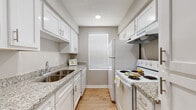 This single-floor residence offers a well-organized layout with consistent finishes throughout. The galley-style kitchen is a standout feature, equipped with white cabinetry, granite-look countertops, a stainless steel sink, a white electric range, and a refrigerator. Upper and lower cabinets provide ample storage, while a window at the end of the kitchen allows natural light to brighten the space. Wood-look flooring runs throughout the kitchen and continues into the main living areas, creating a cohesive and easy-to-maintain aesthetic. The living area is open and airy, featuring neutral gray walls, white baseboards, and a sliding glass door that leads to an outdoor space, allowing for additional natural light. A secondary room off the main area offers flexibility and connects seamlessly to the kitchen vicinity. The bedroom is a comfortable retreat, highlighted by a ceiling fan with light fixture, a window with blinds, and warm wood-look flooring. The bathroom is cleanly appointed with a granite-topped vanity, a toilet, and a shower-tub combination. Neutral tones and consistent flooring throughout lend a unified, modern feel to the entire residence.