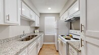 This single-floor residence offers a well-organized layout with consistent finishes throughout. The galley-style kitchen is a standout feature, equipped with white cabinetry, granite-look countertops, a stainless steel sink, a white electric range, and a refrigerator. Upper and lower cabinets provide ample storage, while a window at the end of the kitchen allows natural light to brighten the space. Wood-look flooring runs throughout the kitchen and continues into the main living areas, creating a cohesive and easy-to-maintain aesthetic. The living area is open and airy, featuring neutral gray walls, white baseboards, and a sliding glass door that leads to an outdoor space, allowing for additional natural light. A secondary room off the main area offers flexibility and connects seamlessly to the kitchen vicinity. The bedroom is a comfortable retreat, highlighted by a ceiling fan with light fixture, a window with blinds, and warm wood-look flooring. The bathroom is cleanly appointed with a granite-topped vanity, a toilet, and a shower-tub combination. Neutral tones and consistent flooring throughout lend a unified, modern feel to the entire residence.