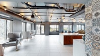This single-floor space showcases a contemporary industrial design that seamlessly blends functionality with sophisticated style. Entry is made through striking steel-framed glass double doors, flanked by decorative mosaic-patterned tile walls that set an artistic tone from the moment of arrival.

The interior opens into a generously proportioned open-concept layout finished with large-format light porcelain tile flooring throughout. Exposed ductwork, conduit, and track lighting run across the dark painted ceiling, lending an industrial character while providing layered illumination across the space.

A central kitchen and bar area serves as the focal point, featuring an extended quartz-topped island with leather bar stools, warm wood cabinetry, and a full-length back counter with a sink and appliances. A bold mosaic tile backsplash and wall-mounted display screen complete this zone. Adjacent to the kitchen is a formal dining area with a dark table surrounded by teal velvet chairs, positioned near floor-to-ceiling windows and sliding doors that open to an outdoor terrace with city views.

A separate lounge nook provides a relaxed retreat with curved grey upholstered armchairs arranged around a sculptural wooden side table. Throughout, large windows flood the space with natural light and frame open urban vistas.