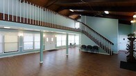This two-story commercial space makes a strong first impression with its generous, open main floor and dramatic architectural character. The ground level features rich, dark hardwood flooring that extends throughout, complemented by soft sage-green walls that lend the interior a calm, welcoming atmosphere. A set of double entry doors with sidelights and flanking windows floods the space with natural light, creating an airy and inviting entryway. Structural columns in a matching green tone add definition to the open floor plan while preserving a sense of flow and openness. A seating area with chairs near the entrance suggests a comfortable waiting or reception zone. The ceiling rises to an impressive vaulted height, finished with warm, dark wood paneling that adds rustic character and visual depth. A ceiling fan helps circulate air throughout the expansive volume. A sweeping curved staircase with dark wood railings and white balusters elegantly connects the main floor to the upper level. The second floor features an open mezzanine with matching railing details that overlooks the main space below, providing excellent sightlines and additional usable area. Together, the two levels create a versatile, well-appointed space suited for a wide range of professional or community-oriented uses.
