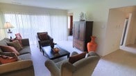 This single-floor space centers around a well-appointed living area that combines comfort and functionality. The room features wall-to-wall carpeting in a neutral tone, contributing to a warm and inviting ambiance throughout. Large windows dressed with vertical blinds span a significant portion of one wall, allowing generous natural light to fill the space while offering privacy control. The seating arrangement includes a dark upholstered armchair accented with rust-colored throw pillows and a pair of generously sized neutral-toned lounge chairs, all oriented around a dark wood rectangular coffee table. A substantial dark wood armoire serves as a prominent storage and display piece along one wall, topped with decorative accents including a sculptural figurine. Vibrant orange accent pieces, including a striking large ceramic vase, introduce bold color contrast against the subdued wall tones and neutral carpeting. A glimpse into an adjacent room reveals continuity in the warm color palette. The overall design aesthetic is traditional and relaxed, combining classic wood furniture with plush seating to create a livable and harmonious environment.