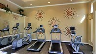 This single-floor fitness room is designed to provide residents or guests with a dedicated space for cardiovascular exercise. The room is equipped with multiple treadmills, each featuring individual monitor screens for entertainment or workout tracking, as well as additional cardio machines positioned throughout the space. The layout is straightforward, allowing users to move comfortably between equipment.

The walls are painted in a warm beige tone and feature a distinctive decorative motif of circular, starburst-style dot patterns in red and dark brown, adding a lively visual element to the space. Recessed lighting in the ceiling provides even, functional illumination throughout the room. The flooring is carpeted, which helps absorb sound and provides a degree of comfort underfoot.

Crown molding along the upper perimeter of the walls adds a subtle architectural detail, lending a slightly more refined finish to what is otherwise a practical, utility-focused space. Overall, the fitness room balances functionality with tasteful design elements, creating an inviting environment for everyday workouts.