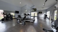 This modern fitness center is designed to support a comprehensive cardio-focused workout experience. The space features soaring vaulted ceilings that create an open, airy atmosphere, making the room feel expansive and energizing. Natural light streams in through large windows, brightening the workout environment and offering a connection to the outdoors.

The facility is outfitted with a variety of cardio equipment, including recumbent bikes, upright bikes, treadmills, and elliptical machines, arranged to allow ample space between each unit for comfortable use. Multiple flat-screen televisions are mounted on the walls, providing entertainment and keeping members engaged during workouts. A large nature-themed artwork is displayed prominently on the upper wall, adding a visual focal point to the space.

The flooring consists of durable, speckled rubberized material well-suited for a high-activity environment. Floor-to-ceiling mirrors line one wall, enhancing the sense of depth and allowing users to monitor their form. Recessed and track lighting supplements the natural light, ensuring the space remains well-lit throughout the day. The overall aesthetic is clean, neutral, and contemporary, with light gray walls contributing to a calm and focused workout atmosphere.