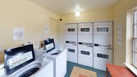 Village Laundry is a practical, single-floor communal laundry facility designed to serve shared-use needs efficiently. The space features warm yellow-toned walls and a clean, functional layout that makes navigation straightforward. The main laundry area is well-equipped with multiple top-loading washing machines and a bank of stacked dryers, providing ample capacity for simultaneous loads. Overhead globe lighting keeps the space bright, while windows along the walls allow natural light to filter in, contributing to an open and airy atmosphere. The flooring features a durable, easy-to-clean surface with a distinctive patterned tile inlay near the entry area, adding a subtle decorative touch. A fire safety pull station is prominently mounted on the wall, reflecting attention to safety compliance. An additional room adjacent to the main laundry area offers extra utility space, complete with a window and standard electrical fixtures. Entry doors provide clear access points, and informational signage is posted throughout to guide users. Overall, the facility is straightforward and well-maintained, offering a comfortable and functional environment for laundry needs.