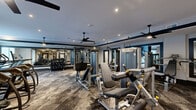 Located at 2050 Stanford Ln in Newport News, VA, this fitness center offers a spacious, single-floor exercise facility designed with both functionality and modern aesthetics in mind. The room is finished with a sophisticated palette of gray and charcoal tones, accented by dark trim molding that frames the walls and ceiling, lending a polished, contemporary feel to the space.

The exercise room is comprehensively outfitted with a wide array of Matrix-brand strength and cardio equipment, including cable machines, weight benches, a full dumbbell rack, treadmills, ellipticals, stationary bikes, and a walking treadmill desk. Full-length mirrors line portions of the walls, amplifying the sense of space and supporting proper form during workouts. Large windows along one wall allow natural light to filter in, creating a bright and energizing atmosphere.

Underfoot, a durable patterned carpet tile in muted earth tones provides both comfort and visual texture. Multiple ceiling fans are suspended throughout the room, ensuring consistent airflow during use. Recessed lighting complements the natural light, keeping the space well-illuminated at all times. A built-in shelving and storage unit near the entrance adds practical organization to the room's refined design, rounding out a fitness facility that is as thoughtfully designed as it is fully equipped.