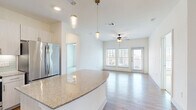 This well-designed single-floor residence offers a bright, open-concept layout that seamlessly connects the kitchen, dining, and living areas. The kitchen is a standout feature, outfitted with sleek light-wood cabinetry, a soft aqua glass tile backsplash, granite countertops, and a full suite of stainless steel appliances including a French door refrigerator, electric range, built-in microwave, and dishwasher. A curved granite-topped island provides additional prep space and casual seating, while elegant pendant lights with brass accents hang overhead. The open living area is bathed in natural light from large windows and a glass door, and is finished with warm light wood flooring. A ceiling fan adds to the comfort of the space. The bedrooms are carpeted for warmth and comfort, each featuring windows with blinds that allow for natural light and privacy. One bedroom includes a sliding glass door that opens to the exterior. The bathroom is finished with matching granite countertops, a tile-surround tub and shower combination, light wood-tone cabinetry, and a large mirror. Throughout the space, neutral white walls, clean lines, and consistent finishes create a cohesive, contemporary aesthetic that is both functional and inviting.