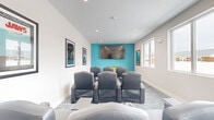 Located at 38 Westgate Dr. in Dover, NH, this single-floor screening and lounge room offers a well-appointed, cinema-inspired experience in a compact yet efficiently designed space. The room is anchored by a bold teal accent wall that frames a large wall-mounted flat-screen television, creating an immediate focal point upon entry. Rows of tufted, dark gray leather recliner chairs are arranged in a theater-style layout, providing ample seating for group viewing. A warm wood-toned credenza beneath the screen adds a touch of natural contrast to the cool-toned palette of soft white and gray that dominates the walls and patterned carpet flooring. Recessed ceiling lighting provides even illumination, while a wall-mounted climate control unit ensures year-round comfort. A series of large windows along the side wall floods the interior with natural light and offers views of the surrounding neighborhood. Classic framed movie posters — including recognizable titles — adorn the walls, reinforcing the cinematic theme and adding personality to the space. The overall ambiance is modern and inviting, blending the comfort of a residential lounge with the dedicated functionality of a private screening room.