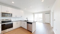 Located at 1636–1648 NW 38th St in Kansas City, MO, this single-floor unit showcases a contemporary design with a cohesive neutral palette and quality finishes throughout. The open-concept layout connects the kitchen and living areas seamlessly, creating a spacious and functional flow.

The kitchen is a standout feature, equipped with white shaker-style cabinetry extending to the ceiling, dark countertops, a white subway tile backsplash, and a full suite of stainless steel appliances including a refrigerator, electric range, microwave, and dishwasher. A peninsula with a built-in sink adds additional prep space and defines the transition into the living area.

The living space is bright and airy, benefiting from large sliding glass doors that open to a private balcony offering open views of the surrounding neighborhood. Light-toned hardwood-style flooring runs throughout the main living areas, enhancing the sense of openness.

The bedroom features plush carpeting, a window with a roller shade for natural light control, and direct access to a bathroom. The bathroom includes a tub-shower combination with white tile surround, wood-tone flooring, a white vanity, and dark countertops, maintaining the unit's consistent modern aesthetic.