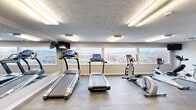 This fitness center offers a comprehensive workout environment housed within a single-level space with a functional, no-frills layout designed to maximize usability. The room features a textured ceiling with flush-mounted fluorescent lighting that evenly illuminates the entire area, creating a bright and energetic atmosphere conducive to exercise.

A bank of large horizontal windows along one wall floods the space with natural light and provides views of the surrounding neighborhood, making cardio sessions more engaging. Multiple treadmills, an elliptical trainer, and a stationary cycle are positioned to take full advantage of these outdoor views, while a wall-mounted television provides additional entertainment options.

Full-length mirrors line one wall, visually expanding the space and allowing users to monitor their form during workouts. The free weight section is well-stocked with a tiered dumbbell rack, medicine balls, and a weight bench, catering to a range of strength training needs. A dedicated stretch and functional training area features a stability dome and open floor space. The durable concrete flooring, partially covered with interlocking rubber mats, provides both practicality and comfort. A structural column near the entry and a glazed entry door with stairwell access complete the room's layout, which also includes a waste station and posted facility guidelines.