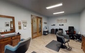 This single-floor hair salon offers a clean, functional, and welcoming environment designed to accommodate a range of beauty services. The space is finished with light hardwood flooring throughout, soft gray walls, and flush panel ceilings with recessed linear lighting that ensures bright, even illumination across all workstations.

The main service area is thoughtfully organized with multiple professional styling stations, each equipped with ergonomic hydraulic chairs, anti-fatigue mats, and warm-toned wooden vanity cabinets topped with framed mirrors. A wall-mounted mini-split climate control unit ensures year-round comfort for both clients and stylists.

A dedicated shampoo station features a reclined wash chair paired with a sleek basin, positioned alongside mobile styling carts stocked with professional tools and equipment. A massage-style pedicure chair rounds out the service offerings, adding a spa-like element to the space.

Rustic wood-trimmed double glass doors with etched detailing serve as a stylish divider between areas, adding a charming design focal point. Natural light filters in through a wood-framed window, offering views of the surrounding outdoor scenery. Decorative wall art and subtle ornamental accents contribute to the salon's warm, inviting ambiance, striking a balance between professional utility and aesthetic appeal.