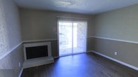 This single-floor residence offers a straightforward and functional layout designed for comfortable everyday living. The living area serves as the centerpiece of the space, highlighted by a classic fireplace with a white surround that adds a traditional focal point to the room. Dark hardwood-style flooring runs throughout, providing a rich, grounded contrast to the neutral-toned walls, which feature a subtle two-tone treatment divided by a chair rail. A textured popcorn ceiling adds a characteristic detail overhead, while crisp white baseboards frame the perimeter of the room. Natural light enters generously through a large sliding glass door fitted with vertical blinds, which opens directly to an outdoor area, offering a seamless connection between the interior and exterior. The sliding door provides views of a brick exterior wall and surrounding greenery, giving the space a sense of openness despite its compact footprint. The overall ambiance is quiet and neutral, presenting a clean canvas well-suited for personalization.