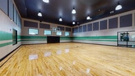 This indoor basketball court is a well-appointed athletic facility designed for serious recreational use. The centerpiece is a gleaming, regulation-size hardwood maple floor, finished to a high polish and marked with standard court lines, including a painted three-point arc and center circle. A glass-backboard basketball hoop is mounted at one end, accompanied by padded wall protection beneath it for player safety.

The walls feature a distinctive design: a bold green and white horizontal stripe runs along the mid-wall perimeter, while large gray acoustic panels are mounted near the ceiling to manage sound within the space. The dark ceiling is elevated well above the playing surface, providing generous vertical clearance appropriate for competitive play, and is fitted with industrial-style pendant light fixtures that evenly illuminate the court below.

Clerestory-style windows are positioned along multiple walls, allowing views into an adjacent fitness and equipment room, which is visible through large interior windows and glass-panel doors. This connectivity suggests the court is part of a broader athletic complex. Entry and exit are provided via multiple black-framed glass doors along one wall. The overall ambiance is clean, functional, and purpose-built, combining practical athletic infrastructure with a polished, professional aesthetic.