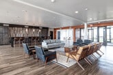Located at 825 Bluegrass Circle in Cedar Falls, Iowa, this contemporary community space offers a thoughtfully designed, open-plan interior spread across a single, generously sized floor. The space makes a strong first impression with its dramatic floor-to-ceiling glass entryway framed in sleek black steel, flooding the interior with natural light and providing clear sightlines to the outdoors.

Throughout the space, warm wood-look luxury vinyl flooring ties together distinct functional zones. The main lounge area is anchored by a modern linear fireplace set against a striking tiled accent wall, complemented by mid-century modern seating arrangements that include sofas, leather chairs, and wooden accent chairs. Bold terracotta-orange accent walls add vibrancy and visual definition between zones.

A casual dining and bar area features dark wood cabinetry, a mosaic tile backsplash, and counter seating, alongside an open dining section with ample table space. A high-ceiling nook bathed in natural light from oversized windows offers bistro-style high-top seating with gold-toned furniture, creating an inviting breakout area.

Structural columns and an open staircase contribute architectural interest, while recessed lighting throughout ensures a warm, well-lit ambiance. The overall design balances comfort and contemporary style, making it a versatile and welcoming communal destination.
