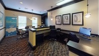 This single-floor professional space is designed to accommodate both collaborative and individual work needs with a polished, business-ready aesthetic. The interior is defined by a striking contrast of cream-toned walls against bold dark trim and crown molding, creating a refined and structured atmosphere throughout.

The workspace is divided into two functional zones. The front area features a built-in workstation counter with a yellow accent base, complemented by a computer desk and printer station. Pendant lighting illuminates this section, while a window allows natural light to filter in, offering views of surrounding greenery.

The conference area centers around a large, dark oval table surrounded by leather executive chairs, making it well-suited for meetings and group discussions. A built-in dark wood bookcase lines one wall, stocked with books and reference materials, adding both utility and visual depth. Additional windows fitted with blinds provide controllable natural light.

Throughout the space, framed artwork in matching dark frames adds a curated, professional touch. The patterned carpet in muted earth tones grounds the design, tying together the dark furnishings and neutral walls into a cohesive, functional environment.