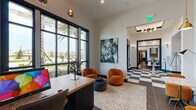 This sophisticated single-floor commercial space makes a striking first impression with its bold black-and-white checkered tile flooring that spans the grand entry vestibule, setting a dramatic tone from the moment of arrival. Floor-to-ceiling black-framed windows flood the interior with abundant natural light while framing outdoor views of palm trees and landscaped surroundings, creating a seamless connection between the interior and exterior environments.

The reception area features a sleek wooden desk paired with a modern computer workstation, complemented by plush burnt-orange lounge chairs and a woven area rug. Statement artwork adorns the walls, adding an artistic, contemporary flair to the professional setting. Pendant lighting with amber-toned glass globes and a warm wood ceiling accent contribute to the inviting ambiance.

A corridor lined with wall sconces leads to a distinctive lounge area, where elegant arched alcoves with soft blue-grey upholstered walls house intimate seating nooks. Each alcove is fitted with a marble-topped bistro table, curved velvet seating, and brass pendant and wall-mounted fixtures, creating a refined yet comfortable atmosphere. Dark large-format tile flooring grounds the lounge, while the tall ceilings throughout enhance the overall sense of openness and sophistication.
