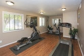 Located at 5222 Wyoming Blvd NE in Albuquerque, New Mexico, this single-floor fitness room is a purpose-built exercise space designed to provide residents with a convenient and well-equipped workout environment. The room features warm hardwood-style flooring throughout, complemented by neutral taupe walls and a textured ceiling with flush-mounted ceiling lights that ensure bright, consistent illumination.

The space is outfitted with three key pieces of cardio equipment: a treadmill, an elliptical trainer, and a stationary recumbent bike, each positioned on protective floor mats. A large decorative mirror with an ornate silver frame is mounted on one wall, visually expanding the room and allowing users to monitor their form during workouts. Colorful abstract wall art adds a touch of personality to the otherwise functional space.

Natural light filters in through multiple windows fitted with white horizontal blinds, including a sidelight flanking a white paneled entry door with an arched transom window. A fire extinguisher is mounted near the entrance, reflecting attention to safety. Ventilation is provided through ceiling and floor-level vents, ensuring a comfortable workout environment. A decorative potted plant adds a subtle organic element, softening the utilitarian nature of the room. Overall, the space is clean, functional, and thoughtfully arranged for everyday fitness use.