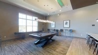 This single-floor game room is designed with a contemporary aesthetic, offering a well-appointed space for social gatherings and leisure activities. The centerpiece of the room is a striking dark-finished billiards table with a bold, sculptural X-shaped base, positioned beneath an elongated, multi-bulb pendant light fixture that provides focused illumination for gameplay.

The ceiling features a coffered tray design with subtle green accent lighting along its edges, adding a modern, upscale touch to the space. Recessed lighting throughout ensures the room remains well-lit, complementing the natural light that streams in through a large multi-pane window along one wall, offering views of the surrounding landscape.

The flooring consists of a striped, multi-tone carpet that adds texture and visual interest while defining the play area. High-top tables paired with sleek, industrial-style bar chairs line the perimeter, providing casual seating for spectators and socializing. Abstract framed artwork adorns the neutral-toned walls, contributing to the room's polished, contemporary character. The overall ambiance is sophisticated yet inviting, purpose-built for recreation and entertainment.
