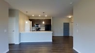 This single-floor space showcases a thoughtfully designed open-concept layout that seamlessly connects the kitchen and living areas. The kitchen is a standout feature, equipped with dark espresso cabinetry that provides ample storage, black appliances including a refrigerator and range, and a spacious peninsula with a light-colored countertop that doubles as a breakfast bar. Two pendant lights hang above the peninsula, adding focused task lighting and a touch of style. Recessed lighting throughout the ceiling supplements the overall brightness of the space.

Rich, dark hardwood-style flooring runs throughout, creating a warm contrast against the neutral grey-toned walls and white trim. The open layout allows for flexible furniture arrangement in the living area, which benefits from the generous flow of the connected spaces. A grey paneled door leads to what appears to be a hallway with additional rooms beyond, hinting at further living space. The overall design balances contemporary aesthetics with functional everyday living, making it a well-appointed and cohesive environment.