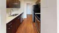 This single-floor unit is finished with rich, dark hardwood flooring that runs consistently throughout every room, creating a cohesive and warm aesthetic. The layout, as seen in the floor plan, includes a spacious living area, a separate bedroom wing, a kitchen, and a full bathroom, all connected by a practical interior corridor.

The living area is generously proportioned, featuring cream-toned walls, clean white baseboards, and a textured ceiling. Natural light enters through a window fitted with horizontal blinds, offering views of surrounding greenery and a distant skyline. A baseboard heating unit is positioned beneath the window for efficient climate control.

The kitchen is designed in a galley configuration, featuring dark espresso cabinetry paired with light countertops and a stainless steel sink. Appliances include a black refrigerator, an electric range, a built-in microwave, and a dishwasher, providing a fully functional cooking space.

The bedroom area is well-sized, benefiting from its own window with outdoor views, and includes multiple closets with bi-fold doors and built-in rods for organized storage. The full bathroom is clean and functional, with a tiled tub-and-shower combo, toilet, and vanity, all complemented by the same hardwood flooring found throughout the unit.