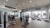 Located at 502 NW 207th St in Miami Gardens, Florida, this single-story fitness center is a comprehensively outfitted facility designed to support a wide range of workout routines. The space is organized into distinct functional zones, each catering to different training disciplines.

The cardio area features a row of modern treadmills and an elliptical machine positioned along a glass door wall, allowing natural light to fill the space and providing views of an outdoor terrace. The strength training section includes a multi-station cable machine, selectorized weight machines, and an adjustable bench, supported by a full rack of dumbbells arranged along floor-to-ceiling mirrored walls that enhance the sense of openness. A dedicated functional training zone houses a power rack with a punching bag, a battle rope, an air bike, and a stability ball, offering versatility for high-intensity and cross-training workouts.

Throughout the facility, bold motivational phrases — "EXECUTION," "GRIT," and "GRIND" — are displayed prominently on the light gray walls, reinforcing an energetic atmosphere. Recessed ceiling lighting provides consistent, even illumination across the space. Durable rubber flooring covers the entire area, ensuring both safety and practicality. Glass double doors open directly to an outdoor patio, connecting the interior workout space to the fresh air outside.