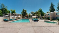 Located at 6312 Birdcage St in Citrus Heights, California, this outdoor amenity space serves as a central gathering hub for the surrounding residential community. The centerpiece is a generously sized rectangular swimming pool with crystal-clear blue water, designed for both leisure and lap swimming. Adjacent to the pool sits a spa, offering a dedicated space for relaxation. The expansive concrete sun deck surrounds the pool on all sides, providing ample room for lounging and socializing. Teal-cushioned chaise lounges are arranged throughout the deck, accompanied by market umbrellas that offer shade during sunny California afternoons. A canopied gazebo structure adds an additional shaded retreat, ideal for more private relaxation. A dedicated seating and dining area with patio furniture provides a comfortable space for gatherings. The pool area is enclosed by a perimeter wall and fencing, ensuring privacy and security. A clubhouse-style building with French doors borders one side of the deck, offering convenient indoor access. Mature trees, including towering pines, frame the space and contribute to a serene, park-like atmosphere. The overall ambiance is bright, open, and inviting — well-suited for outdoor recreation under the Northern California sun.