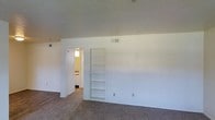 This single-floor space offers a clean, functional layout finished in neutral whites and warm beige tones throughout. The main living area features wall-to-wall carpeting that adds warmth underfoot, while the smooth white walls provide a bright, open atmosphere. A standout architectural detail is the built-in recessed shelving unit integrated directly into the wall, offering practical storage and display space without encroaching on the room's floor area. HVAC vents are thoughtfully positioned both near the ceiling and lower on the walls, ensuring efficient climate control throughout the space. An open doorway leads directly into an adjacent room, which appears to be a bathroom, finished with lighter walls and overhead lighting, making it conveniently accessible from the main living area. The overall design is straightforward and understated, prioritizing functionality with a neutral palette that provides a versatile backdrop for a variety of uses. The single-floor layout ensures easy navigation between spaces, while the consistent use of soft, muted tones creates a cohesive and calm ambiance throughout.
