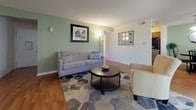 Located at 6741 E Tanners Creek Dr in Norfolk, Virginia, this single-floor unit offers a well-organized layout with comfortable living spaces and practical finishes throughout. The living room features warm wood-style flooring, a distinctive sage green accent wall, and natural light, creating a welcoming and relaxed atmosphere. Adjacent to the living area, the kitchen is equipped with rich wooden cabinetry, dark granite-style countertops, black appliances including a range, dishwasher, and refrigerator, and durable hardwood-style flooring. The dining area, connected to the kitchen, provides additional space for everyday meals and includes a window with views of the surrounding greenery, along with in-unit laundry access. The unit includes two bedrooms, both carpeted for comfort. The first bedroom is generously sized with large windows allowing ample natural light and features a spacious closet with sliding doors. The second bedroom is equally comfortable, with windows and easy access to additional storage. The full bathroom is finished with tile flooring, a combined tub and shower, and a wooden vanity with a white countertop. Throughout the unit, neutral tones and consistent finishes contribute to a cohesive and functional living environment.