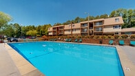 Located at 100 Pinegate Cir in Chapel Hill, North Carolina, this outdoor pool area offers a well-appointed recreational space designed for relaxation and leisure. The centerpiece is a generously sized swimming pool with vivid blue water, lane markers, and a smooth concrete surround that provides ample room for sunbathing and social gathering.

Surrounding the pool, a generous number of teal-colored lounge chairs are arranged along all sides, offering plenty of seating for residents. Several shaded areas are available, with colorful orange umbrellas providing relief from the sun, accompanied by additional table and chair seating for a more casual outdoor dining or lounging experience.

The pool area is enclosed by a sturdy black iron fence, complemented by a well-maintained brick retaining wall with lush landscaping including vibrant greenery and flowering plants. The backdrop features multi-story residential buildings with balconies, framed by mature trees that lend a natural, serene atmosphere to the setting.

Safety features are visible throughout, including pool safety equipment stations. The space benefits from an open-sky orientation, allowing for abundant natural sunlight throughout the day. Overall, the pool area presents a clean, inviting, and well-maintained outdoor amenity space ideal for warm-weather enjoyment in the heart of Chapel Hill.