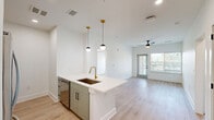 Located in Arden, NC, this thoughtfully designed 2-bedroom, 2-bathroom residence offers a clean, modern aesthetic throughout its single-floor layout. The open-concept kitchen and living area serves as the heart of the home, anchored by a sleek central island with a white quartz countertop and a brushed gold faucet. Light sage cabinetry pairs with stainless steel appliances, while pendant globe lights with gold hardware add a contemporary touch. Light-toned hardwood-style flooring flows throughout the main living areas, complementing the bright white walls and creating a cohesive, airy feel. The living room benefits from natural light through large windows and a glass patio door, with a ceiling fan overhead. The patio provides an outdoor retreat with views of the surrounding greenery. Both bedrooms feature plush carpeting, ceiling fans, and windows that invite natural light. The primary bedroom connects to a well-appointed en-suite bathroom featuring a double-sink vanity with light wood cabinetry, a framed mirror, matte black hardware, and a walk-in glass shower. The second bathroom mirrors this refined style. Additional highlights include four closets offering generous storage, a dedicated laundry space, and two hallways that ensure efficient circulation throughout the home.