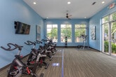 This single-floor fitness center is designed to support a comprehensive workout experience in a bright, welcoming environment. The space is finished with soft blue walls and textured carpet flooring throughout, creating a calm yet energizing atmosphere.

The main exercise area is generously sized and benefits from abundant natural light streaming through large, floor-to-ceiling windows that overlook lush, tree-lined outdoor surroundings. A ceiling fan overhead helps maintain airflow and comfort during workouts. The room accommodates a variety of cardio equipment, including treadmills and a multi-function strength training tower, with open floor space available for stretching or additional exercises.

A dedicated spin studio area features a row of professional-grade stationary cycling bikes, a wall-mounted television for guided sessions, and a stability ball for supplementary training. The studio is clearly defined within the broader space and offers an ideal setup for group or individual cycling workouts.

The entryway area provides a practical transition zone with storage for equipment such as yoga mats and accessories, alongside a digital control panel and a stainless steel waste bin. Large glass entry doors open directly to the outside, reinforcing the connection to the outdoors and allowing for easy access. Motivational artwork is displayed throughout, adding a dynamic visual element to the overall design.