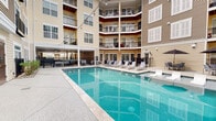 Located at 307 Karl Linn Dr in North Chesterfield, Virginia, this outdoor amenity space is thoughtfully designed to serve as a central gathering hub for its residential community. The centerpiece is a sparkling resort-style pool framed by a generous pool deck lined with lounge chairs, ideal for relaxation and recreation. Surrounding the pool, a spacious covered patio area provides a comfortable retreat from the elements, featuring beadboard ceilings with ceiling fans, wall-mounted televisions, and a mix of sectional seating and dining tables with chairs — all styled in a clean, contemporary palette of white, gray, and blue accents. The covered space transitions seamlessly into an open-air courtyard where a built-in outdoor kitchen with a gas grill and a stone-accented fire pit with Adirondack chairs create an inviting setting for outdoor entertaining. Paved walkways with decorative brick accents, manicured landscaping with shrubs and rock beds, and warm string lights overhead add to the welcoming ambiance. The space is nestled within the inner courtyard of a multi-story building featuring tan and blue siding, wood-detailed balconies, and black railings, creating an enclosed and community-oriented atmosphere throughout.