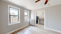 Located at 5541 S Everett Ave in Chicago's Hyde Park neighborhood, this single-floor unit offers a well-organized layout with consistent finishes throughout. Light-toned hardwood flooring runs seamlessly across all rooms, creating a bright, cohesive feel. Crown molding and clean white trim add a classic architectural touch to the neutral gray walls.

The main living area is generously proportioned, featuring a ceiling fan with light fixture and a large sliding closet. Multiple double-hung windows allow abundant natural light and frame views of the surrounding Chicago cityscape. Classic cast-iron radiators provide traditional heating in both the living and secondary rooms.

The secondary room offers a similarly open layout with additional windows and direct access to the compact kitchen. The kitchen is efficiently designed, featuring warm wood cabinetry, a built-in gas range with oven, a single-basin sink, and a countertop workspace — providing all essential cooking amenities in a streamlined footprint.

The bathroom is clean and functional, with a full soaking tub and shower combination, a white vanity with sink, and a window that introduces natural light. Built-in shelving adds practical storage. Overall, the unit presents a tidy, move-in ready space with practical amenities and a classic Chicago residential character.
