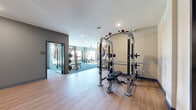 This modern fitness center at 10631 Parallel Pkwy in Kansas City, KS, offers a comprehensive workout environment spread across a generously sized single-floor layout. The facility is divided into distinct functional zones, each thoughtfully designed to support a wide range of fitness activities.

The main strength and conditioning area features light wood-look flooring, neutral gray walls, and recessed lighting. It is outfitted with a multi-functional cable machine, weight benches, plate-loaded equipment, and a dumbbell rack, providing ample options for resistance training. A wall-mounted flat-screen television adds convenience for entertainment during workouts.

A striking double-height training zone commands attention with floor-to-ceiling windows on multiple walls, flooding the space with abundant natural light and offering open views of the surrounding Kansas City skyline. This area features an artificial turf running lane alongside carpeted flooring, battle ropes, and additional strength machines — ideal for functional and athletic training.

Decorative accents elevate the overall aesthetic, including a bold geometric accent wall with a large ceiling fan, vibrant abstract artwork, and colorful murals that give the facility a dynamic, modern character.

A dedicated recovery room houses a wood-paneled infrared sauna with chromotherapy lighting, providing a private wellness retreat within the facility. The adjoining corridor features built-in storage cabinetry and connects the various zones seamlessly.