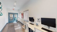 This single-floor amenity space is thoughtfully designed to serve as both a functional business center and a casual social hub. The layout flows seamlessly from a well-equipped workstation area into an open game room, offering a versatile environment for productivity and leisure alike.

The business center section features a long, sleek white desk lined with multiple computer workstations and ergonomic rolling chairs, making it ideal for focused work. The crisp white walls and wood-look flooring contribute to a clean, contemporary aesthetic, while layered zebra-stripe roller blinds on the windows allow for customizable natural light filtering throughout the day.

A cozy lounge nook with a round glass-top table and modern white chairs provides an intimate spot for casual meetings or relaxed reading. A vibrant abstract painting on the wall adds a pop of color and artistic character to the space.

The corridor connects seamlessly to an adjacent game room, where a full-size pool table anchored on a patterned area rug becomes a natural social focal point. A ceiling-mounted pendant light illuminates the game area, adding warmth to the otherwise bright, airy interior.

The entrance features striking teal double doors with glass panels, offering a welcoming and stylish first impression. A patterned runner rug along the hallway adds texture and visual interest, tying together the space's modern, inviting design.