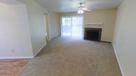 This single-floor space offers a comfortable and functional layout designed for everyday living. The main living area is generously sized, featuring wall-to-wall carpeting that adds warmth and softness underfoot. A standout feature of the room is the classic brick fireplace, complete with a raised hearth and a clean white mantle, serving as a natural focal point. Opposite the fireplace, a large sliding glass door with vertical blinds allows an abundance of natural light to flood the interior, creating a bright and airy atmosphere while providing direct access to an outdoor area. A ceiling fan with an integrated light fixture is centrally positioned overhead, contributing to both comfort and airflow throughout the space. The walls are finished in a neutral, light tone, complemented by a textured ceiling that adds subtle visual interest. A hallway visible from the main living area suggests additional rooms and functional spaces beyond. The overall ambiance is clean, open, and move-in ready, offering a solid foundation for personalization and comfortable day-to-day living.