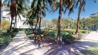 Located at 250 Congress Park Dr in Delray Beach, Florida, this outdoor space offers a beautifully maintained tropical landscape that embodies the character of South Florida's natural environment. A smooth, paved walkway winds through the area, flanked by towering palm trees whose fronds stretch high above, providing natural shade and a distinctly tropical ambiance. Vibrant tropical plantings, including bold bromeliad-like foliage and dense ground-cover shrubs, are thoughtfully arranged throughout the landscape, adding layers of color and texture. Flowering plants punctuate the greenery with splashes of warm color, enhancing the visual appeal of the corridor. The setting is bathed in abundant natural sunlight under a clear blue sky, creating a bright and inviting atmosphere. In the background, multi-story buildings with white and light-colored facades are visible through the tree canopy, indicating that this landscaped pathway serves as an outdoor common area within a larger commercial or mixed-use campus. Low-profile landscape lighting fixtures are positioned along the plantings, suggesting the walkway remains accessible and well-lit in the evening hours. Overall, the space presents a lush, well-curated outdoor environment ideal for pedestrian circulation and casual outdoor enjoyment.