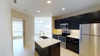 This single-floor space showcases a contemporary kitchen designed with both style and functionality in mind. The kitchen is anchored by a central island topped with a crisp white quartz countertop and an undermount sink, providing ample workspace and a natural focal point for the room. Dark espresso-toned cabinetry lines the walls, offering generous storage while creating a striking contrast against the light-colored countertops and neutral beige walls.

Stainless steel appliances, including a built-in microwave, electric range with oven, and dishwasher, are seamlessly integrated into the cabinetry, lending the space a polished, modern aesthetic. Recessed lighting throughout the ceiling ensures even, bright illumination across the entire kitchen area.

A standout feature of this space is the expansive floor-to-ceiling windows that flood the kitchen with natural light and frame impressive views of the surrounding urban skyline. Light wood-toned flooring runs throughout, complementing the overall design and adding warmth to the space. The open layout connects the kitchen naturally to the adjacent living area, enhancing the sense of spaciousness and flow.