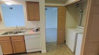 This two-floor space is designed with functionality and practicality at its core. The kitchen is equipped with warm oak cabinetry, a dishwasher, and a sink set into a light-toned countertop, providing ample workspace for meal preparation. The cabinetry extends to the upper walls, offering generous storage options throughout the area. Adjacent to the kitchen, a dedicated laundry space is conveniently tucked in, complete with a washer and dryer unit and an overhead shelf for additional storage — a thoughtful layout that keeps household tasks organized and efficient. The tile flooring in this area is both durable and easy to maintain, complementing the neutral, light-toned walls that give the space a clean, open feel. A doorway connects to a carpeted room beyond, hinting at a seamless flow between the utility areas and the living spaces of the home. The overall design emphasizes a no-fuss, livable environment, with a layout that prioritizes ease of use across both floors of the property.