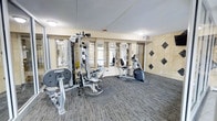 This fitness center is a purpose-built exercise facility designed to support a full range of workout routines. The space is anchored by a multi-station strength training machine by Hoist, offering a variety of resistance-based exercises in a compact footprint. Complementing the strength equipment is a stationary recumbent bike, ideal for low-impact cardiovascular training. The floor is covered in durable, textured carpet tiles that provide both comfort and practicality underfoot. Mirrored wall panels line one side of the room, enhancing the sense of openness and allowing users to monitor their form during exercise. The walls feature an elegant tile treatment, with large neutral-toned panels accented by dark diamond-shaped tile insets, adding a polished, upscale character to the space. Ceiling-mounted flush lights provide consistent, even illumination throughout. An exit door with signage is clearly visible, ensuring safe and convenient access. The overall layout is efficiently organized, making practical use of the available floor area while maintaining clear pathways between equipment. The space conveys a clean, professional atmosphere well-suited for residents or guests seeking a convenient on-site fitness option.