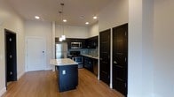 This well-appointed kitchen showcases a cohesive modern design built around striking dark espresso cabinetry that lines the walls and provides generous storage throughout. A central island anchors the space, topped with light-toned stone countertops that offer ample prep surface and create a strong visual contrast against the dark base. Overhead, a pair of sleek pendant lights hang at varying heights, casting focused illumination over the island and adding a contemporary design element.

The kitchen is fully equipped with stainless steel appliances, including a refrigerator, electric range, and built-in microwave, all seamlessly integrated into the cabinetry layout. A tiled backsplash in a neutral mosaic pattern runs along the cooking and sink areas, adding texture while remaining easy to maintain. The sink is positioned beneath upper cabinetry along the perimeter, maximizing functional workspace.

Warm hardwood-style flooring extends throughout, lending a sense of continuity and warmth to the space. Recessed ceiling lights supplement the pendant fixtures, ensuring even illumination. Two dark-paneled doors with sleek hardware are visible, maintaining the space's modern aesthetic and providing access to adjacent areas.