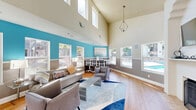 Located at 2850 Middlefield Rd in Palo Alto, California, this well-appointed community living room offers a welcoming and versatile gathering space. The room immediately impresses with its soaring vaulted ceilings, which lend an open, airy feel that extends well beyond its compact footprint. Warm hardwood floors run throughout, complemented by a two-tone wall treatment — bold teal on the upper half and neutral taupe below — that gives the space a modern yet inviting character.

Natural light pours in through multiple large windows that frame views of the surrounding landscaping and a nearby pool, creating a bright and cheerful atmosphere throughout the day. A classic fireplace with a built-in mantle serves as a focal point on one wall, paired with a wall-mounted flat-screen television above it for added functionality.

The living area is thoughtfully furnished with a contemporary sofa, lounge chairs, and a geometric blue-and-white area rug that anchors the seating arrangement. A dedicated workspace with a desk station and ergonomic chairs provides a practical area for residents. A casual dining high-top table with stools near the window adds a social element to the layout. The entryway features double glass doors and French glass panel doors, connecting the space seamlessly to the outdoors and an adjacent area.