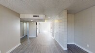 This single-floor residence offers a practical and functional layout across two bedrooms, one bathroom, and a combined kitchen and living area. Throughout the space, light-toned wood-look flooring provides a cohesive aesthetic, complemented by textured ceilings and neutral warm-gray walls that create a clean, understated ambiance.

The open kitchen and living area is anchored by a distinctive white tile-block accent wall that extends from floor to ceiling, adding architectural character to the space. A stainless steel refrigerator is integrated into the kitchen area, and large vertical-blind-covered windows allow abundant natural light to fill the room.

The primary bedroom is a well-proportioned retreat featuring a built-in double closet with sliding doors offering ample storage. The second bedroom is similarly bright, with a window overlooking the outdoors and clean, neutral finishes throughout. Both rooms share the same consistent flooring and textured ceiling treatment.

The bathroom is neatly appointed with a granite-style countertop, an undermount sink with brushed-gold fixtures, a wall-mounted mirror, a soaking tub, and a window that brings in natural light.

A standout functional feature is the in-unit stacked washer and dryer, tucked into a dedicated built-in nook. Central air conditioning with ceiling vents ensures year-round comfort throughout the home.