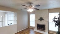 This single-story space centers around a welcoming living room that combines classic and practical design elements. The room features a whitewashed brick fireplace as its focal point, complete with a raised hearth and traditional metal fireplace screen, adding warmth and character to the space. Dark hardwood floors run throughout, providing a rich contrast against the neutral gray walls and offering durability and visual appeal.

A five-blade ceiling fan with an integrated light fixture is centrally mounted on the textured popcorn ceiling, ensuring air circulation and supplemental lighting throughout the room. Multiple windows allow natural light to filter into the space, including a larger window adjacent to the fireplace and a divided-light window on the opposite wall, contributing to a bright and airy atmosphere.

The layout appears open and versatile, suitable for various furniture arrangements. Subtle architectural details such as white baseboards and clean wall transitions add a finished, cohesive look. Overall, the space presents a comfortable, functional living environment with timeless design touches that balance practicality with a lived-in charm.