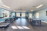This fitness center in Malvern, PA offers a well-appointed, single-floor layout designed to support a range of exercise and recreational activities. The main workout area is spacious and open, finished with durable rubber sport flooring and bathed in natural light from multiple large windows along the perimeter walls. The walls are painted in a calming blue-gray tone, complemented by crisp white crown molding and white trim throughout, lending the space a clean, polished aesthetic. Ceiling fans provide additional air circulation, contributing to a comfortable workout environment. The cardio equipment area features treadmills, elliptical machines, and a stationary bike, each positioned near windows offering pleasant outdoor views. A dedicated free weights section includes a full dumbbell rack with a large wall mirror, ideal for strength training. The open floor plan also accommodates a full-size JOOLA ping-pong table, making it a versatile space for both fitness and recreation. Wall-mounted flat-screen televisions are positioned throughout the room for entertainment during workouts. A bulletin board and informational displays are also present. The facility is completed by a bathroom, ensuring full convenience for users. Accessed via glass double doors, this fitness center is a comprehensive and inviting community amenity.