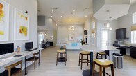This single-story clubhouse offers a thoughtfully designed open-concept interior that combines functionality with contemporary style. The space is characterized by notably high ceilings that enhance the sense of openness and airiness throughout. Light-toned hardwood-style flooring runs throughout the main area, complementing the neutral white and gray color palette of the walls and architectural elements.

The main gathering area features a mix of high-top tables with yellow upholstered bar stools and a comfortable lounge seating arrangement centered around a fireplace, creating distinct zones for both casual socializing and relaxed conversation. A modern sputnik-style chandelier serves as a focal point overhead, adding a decorative touch to the space.

Recessed lighting is distributed across the ceiling to ensure consistent, well-lit conditions throughout. Natural light filters in through multiple windows, contributing to the bright and inviting ambiance. A dedicated workstation area with computer terminals is visible toward the back, offering a practical workspace for residents. A wall-mounted television near the fireplace adds to the entertainment options available in this versatile community gathering space.