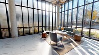 This contemporary lobby makes an immediate impression with its dramatic floor-to-ceiling curtain wall windows framed in sleek black steel, flooding the interior with abundant natural light and offering open views of the surrounding streetscape. The soaring ceiling height amplifies the sense of grandeur and openness throughout the space. Underfoot, large-format marble-look porcelain tile extends across the primary entry and transition areas, while warm hardwood flooring defines the comfortable lounge seating area. A curated mid-century modern seating arrangement anchors the lounge, featuring a tufted teal sofa accented with mustard yellow pillows, upholstered accent chairs, an oval coffee table, and geometric copper side tables, all set atop a layered area rug. A striking starburst chandelier overhead adds a sculptural focal point. Exposed concrete columns punctuate the space, lending an industrial contrast to the refined finishes. A bold deep teal accent wall near the entrance frames glass vestibule doors and provides a vibrant backdrop for curated wall art. The elevator bank is encased in a textured concrete surround, complemented by a sleek console table with decorative accessories. The overall ambiance is modern, airy, and thoughtfully designed, balancing warmth and sophistication.